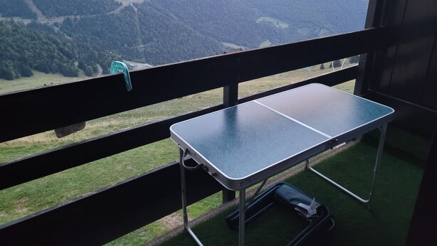 Un balcón con mesa y silla de metal, con vistas a un paisaje montañoso y un cielo despejado.