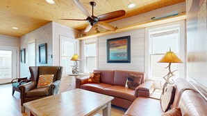 A spacious living room with a ceiling fan, wooden walls, a large window, and a painting. - OV10 Golf Cart Included, Shared Pool, Pet Friendly, Parking for 3 (Port Aransas)