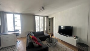 A modern living room with a sofa, a coffee table, and a TV. There is a window with curtains, a radiator, and a door leading to another room. - Saint Honoré - Luxury Apartement 1 BR (Paris)