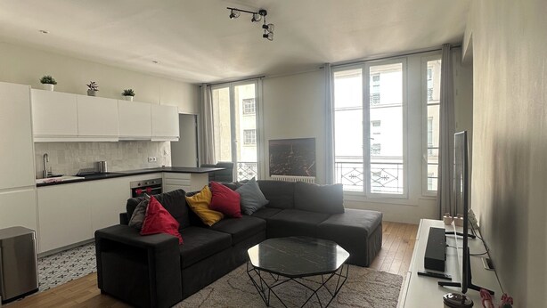 A modern living room with a sectional sofa, a coffee table, and a TV. There is a kitchen area with white cabinets and a countertop. - Saint Honoré - Luxury Apartement 1 BR (Paris)