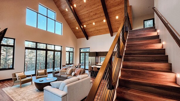 A spacious living room with a high ceiling, large windows, and a wooden staircase.