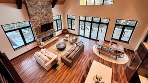 A spacious living room with a stone fireplace, large windows, and a high ceiling.