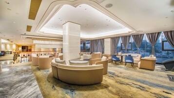 A spacious lobby with a central seating area, a high ceiling with recessed lighting, and large windows offering a view of the city.