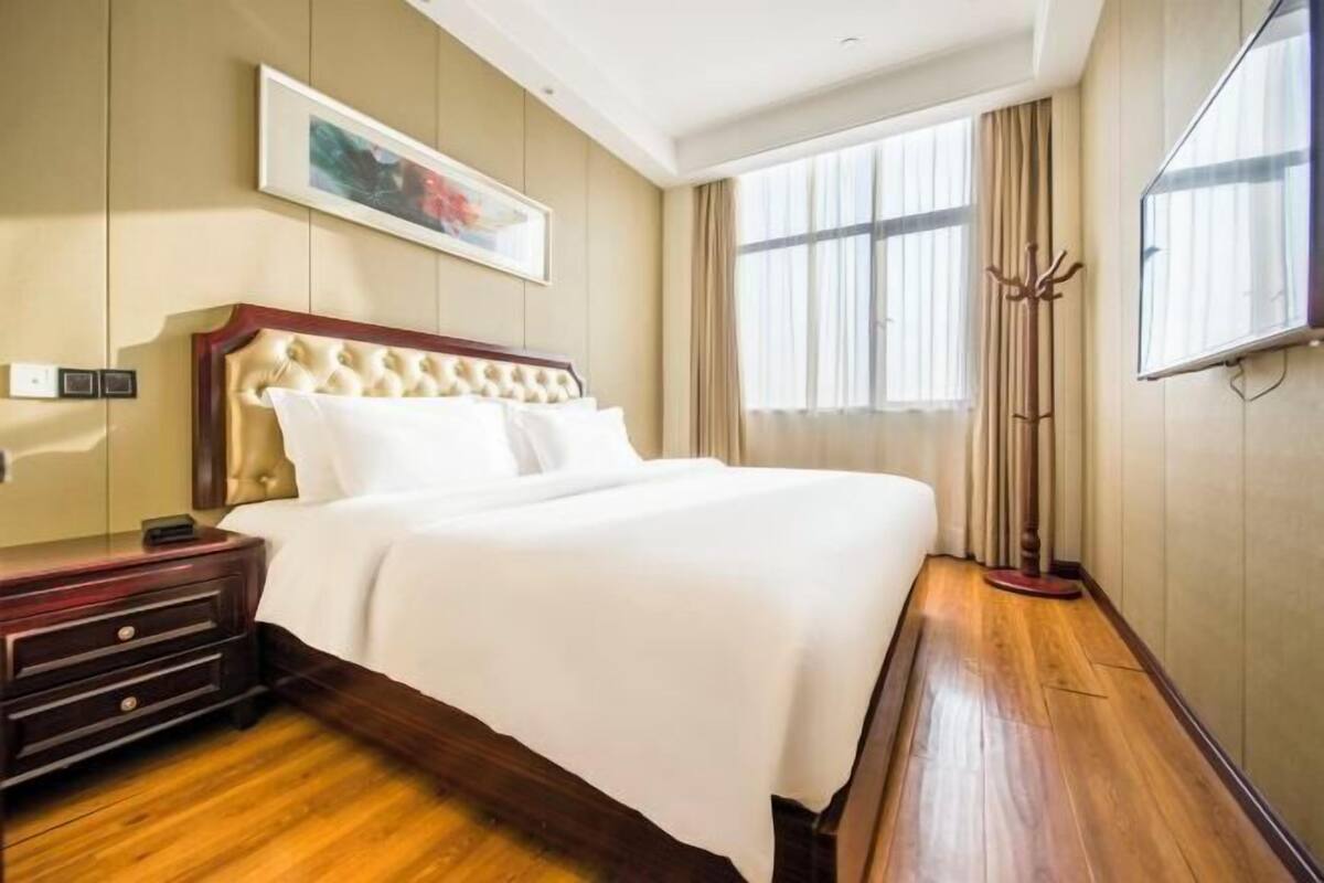 A hotel room with a large bed, a wooden nightstand, a framed picture on the wall, and a large window with curtains.