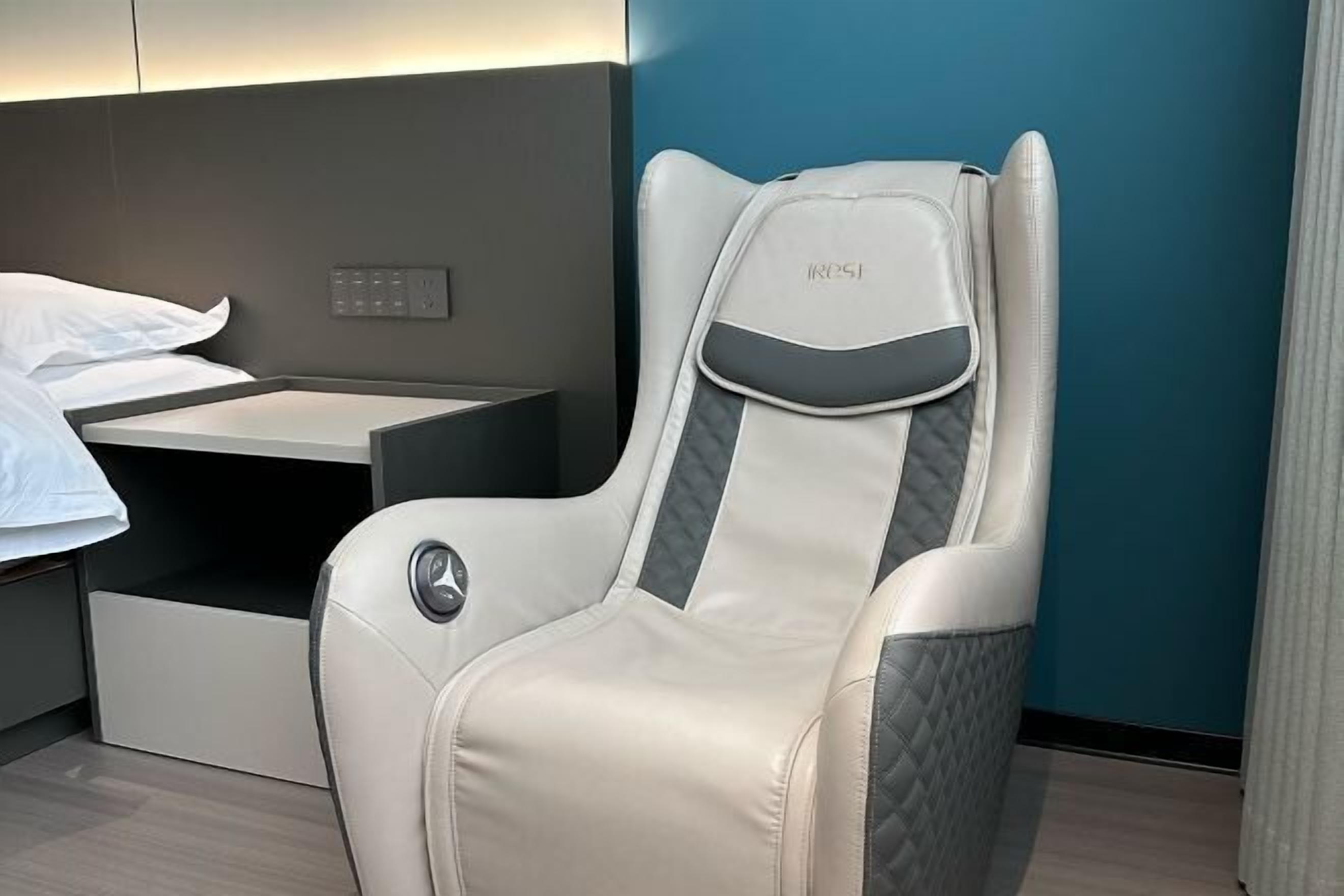 A modern hotel room with a single bed, a bedside table, and a reclining chair with a headrest.