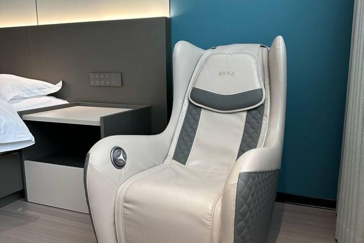 A modern hotel room with a single bed, a bedside table, and a reclining chair with a headrest.