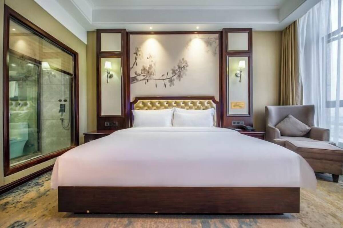 A large bed with a tufted headboard, a glass shower enclosure, and a seating area with a chair.