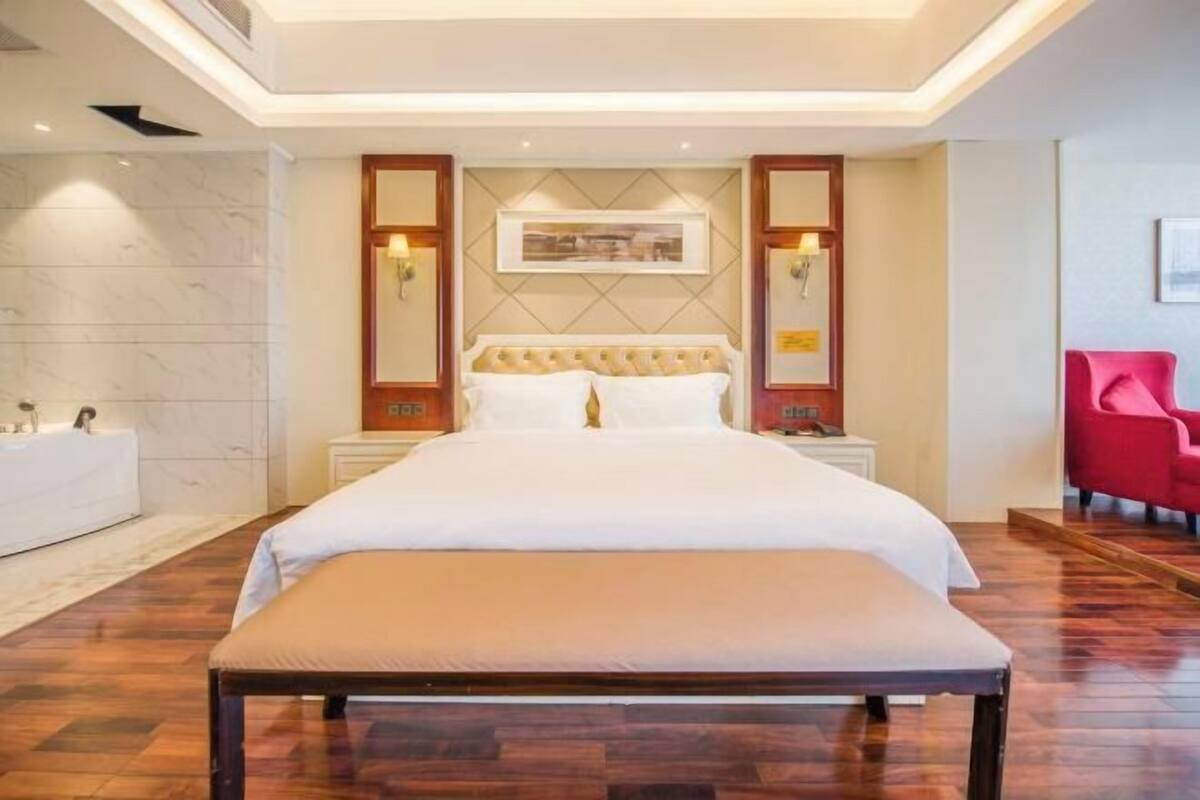 A modern hotel room with a large bed, a bench, a red chair, and a wall-mounted picture.