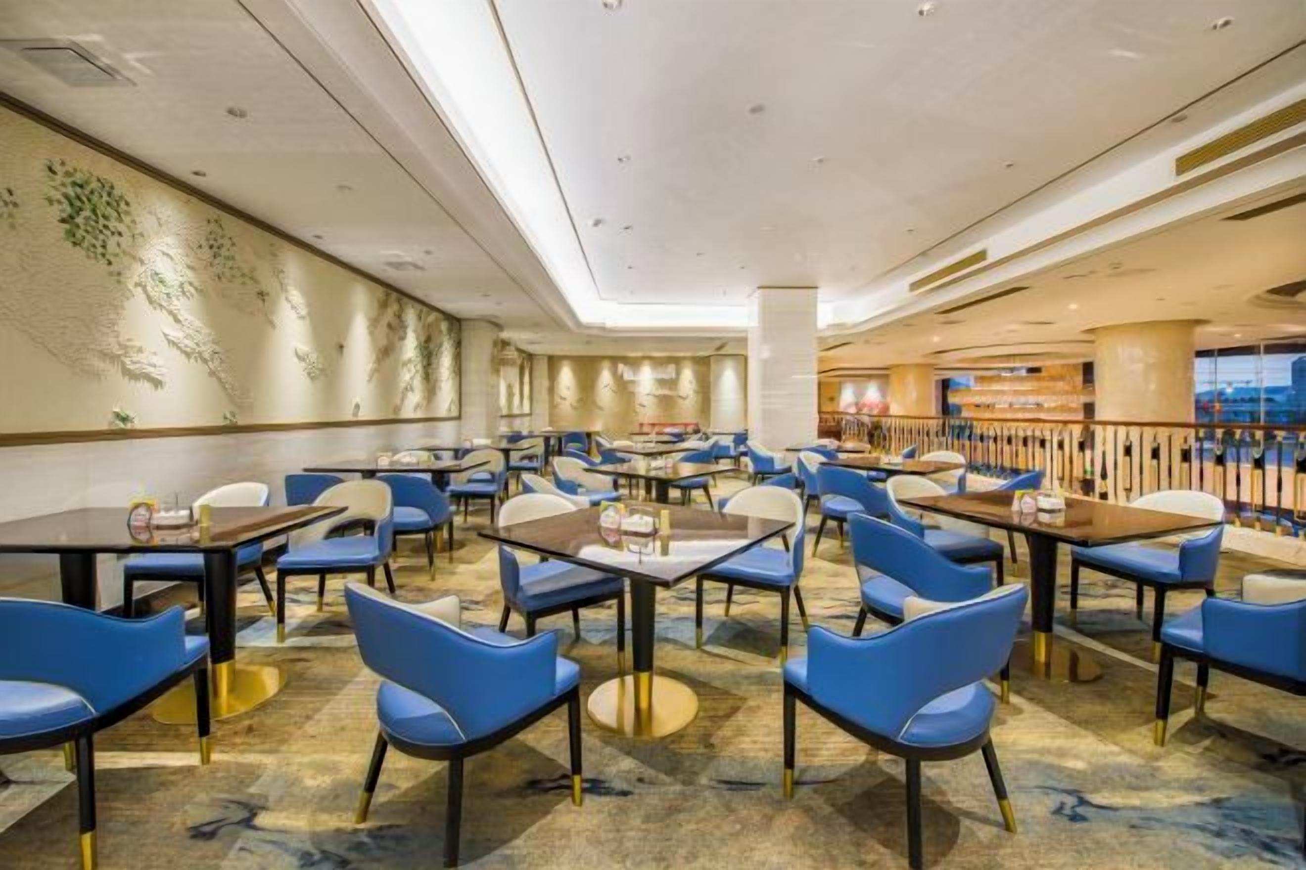 A hotel dining area with multiple tables and chairs, a high ceiling with recessed lighting, and a view of the cityscape through large windows.