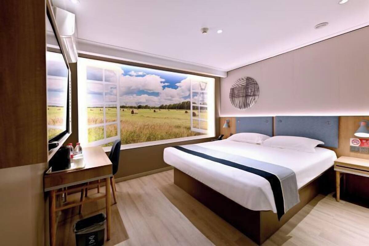 A bedroom with a bed, a desk, a chair, a window with a view of a field, and a wall-mounted light fixture.