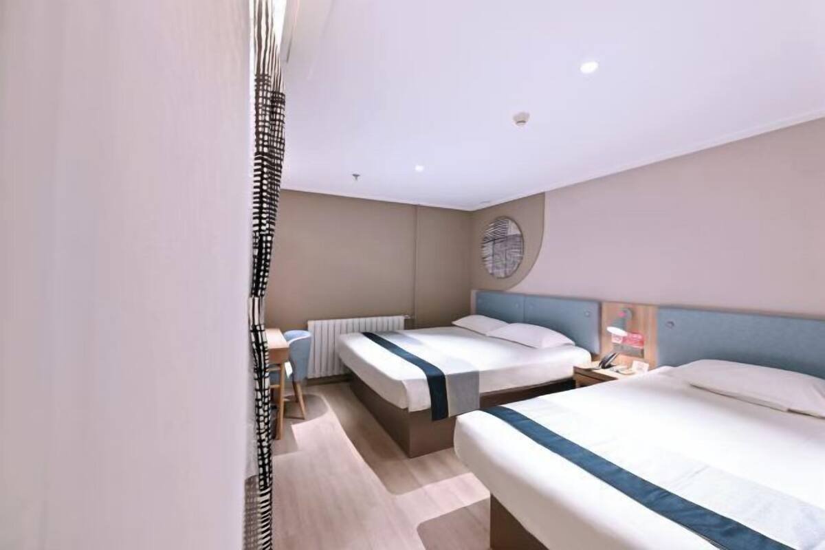 A hotel room with two beds, a desk, a chair, a round mirror, and a radiator.