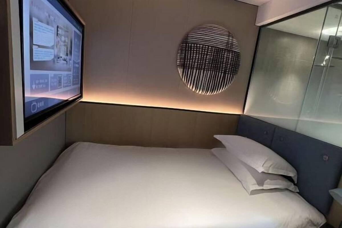 A modern hotel room with a bed, a wall-mounted screen displaying information, and a circular light fixture.