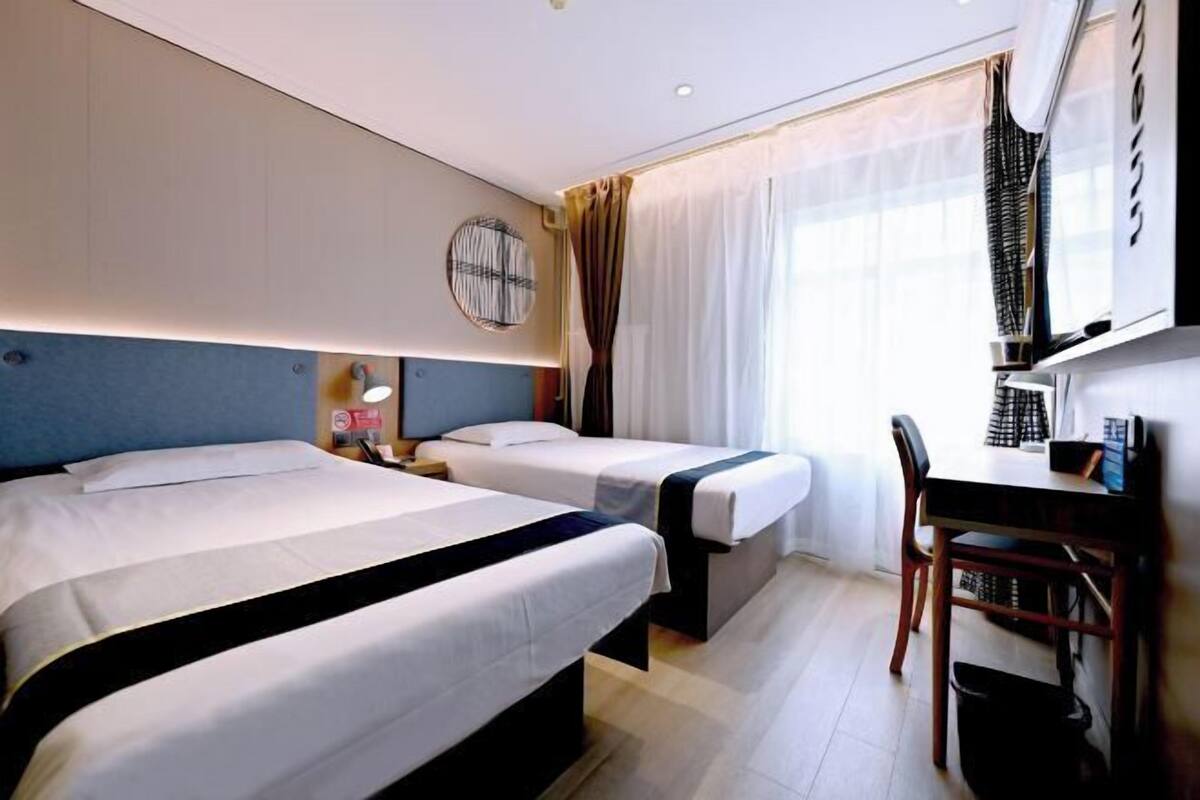 A hotel room with two beds, a desk, a chair, a round wall clock, and a window with curtains.