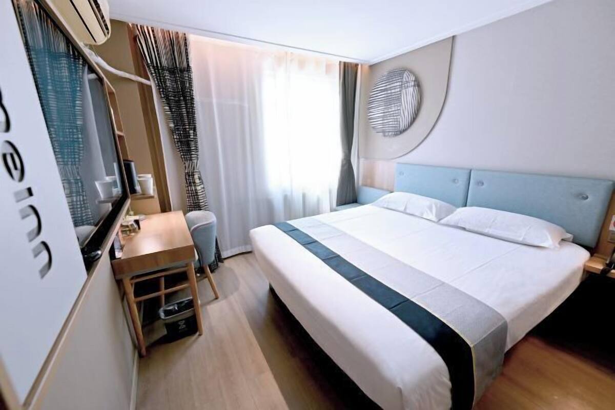 A hotel room with a bed, a desk with a chair, a small table, a wardrobe, and a window with curtains.