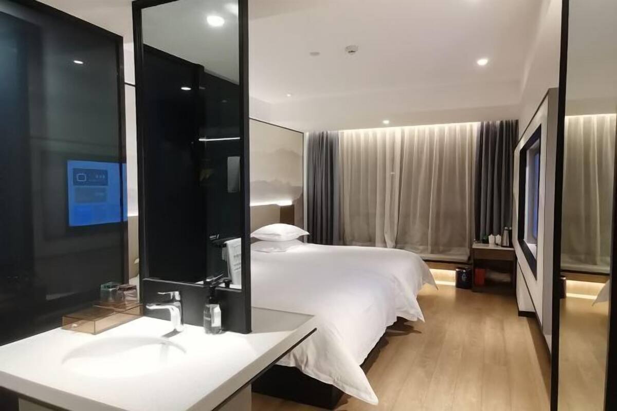 A modern hotel room with a large mirror, a white bed, a bedside table, and a large window with curtains.
