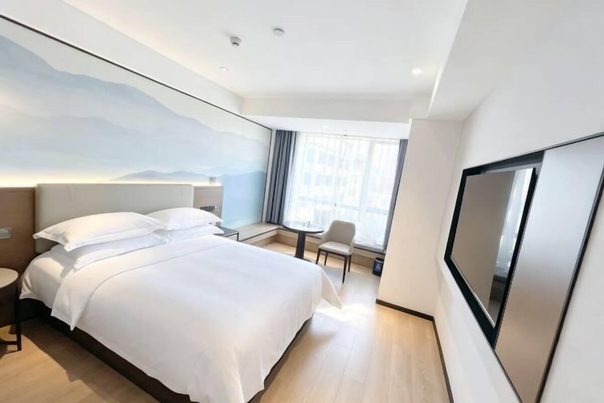 A modern hotel room with a large bed, a desk, a chair, a TV, and a window with curtains.