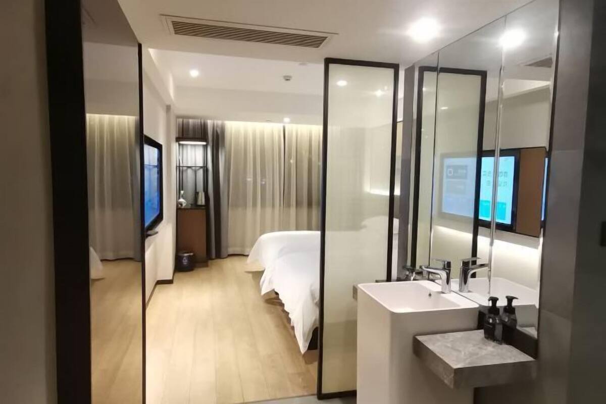 A hotel room with a large mirror, a bed, a bedside table, a TV, and a bathroom with a sink and mirror.