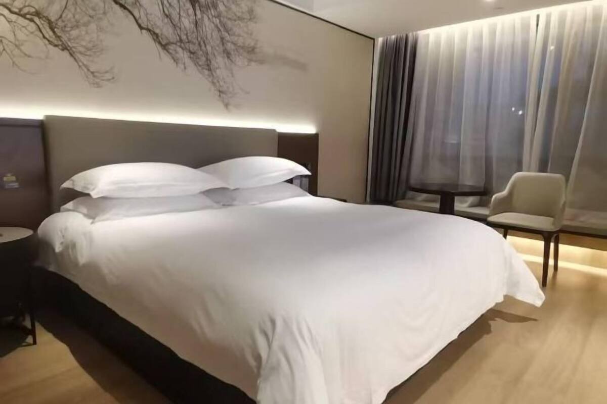 A modern hotel room with a large bed, a bedside table, a chair, and a large window with sheer curtains.