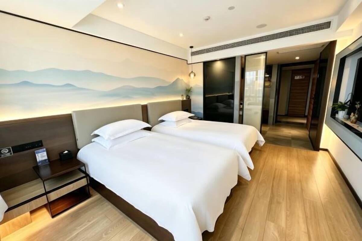 A hotel room with two beds, a nightstand, a wall-mounted TV, and a view of a mountain landscape.