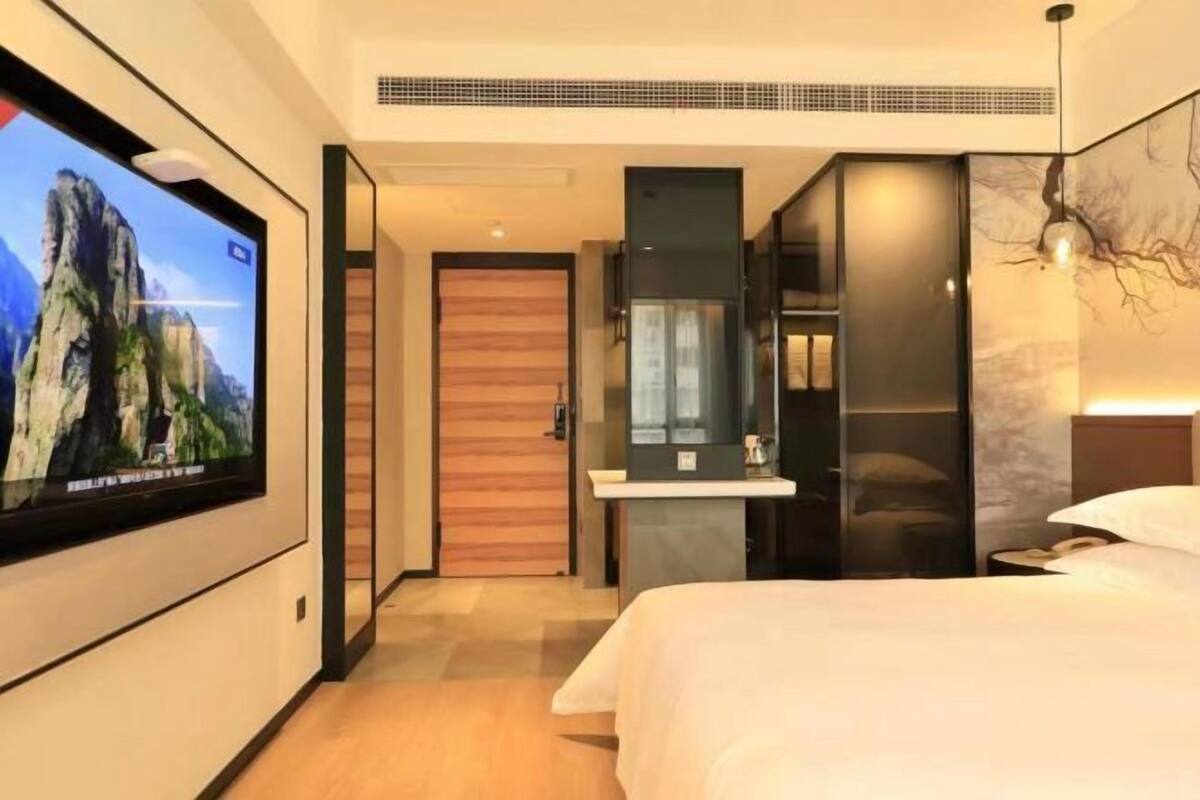 A modern hotel room with a large flat-screen TV mounted on the wall, a bed with white bedding, a wooden door, a small bedside table, and a mirror above it.