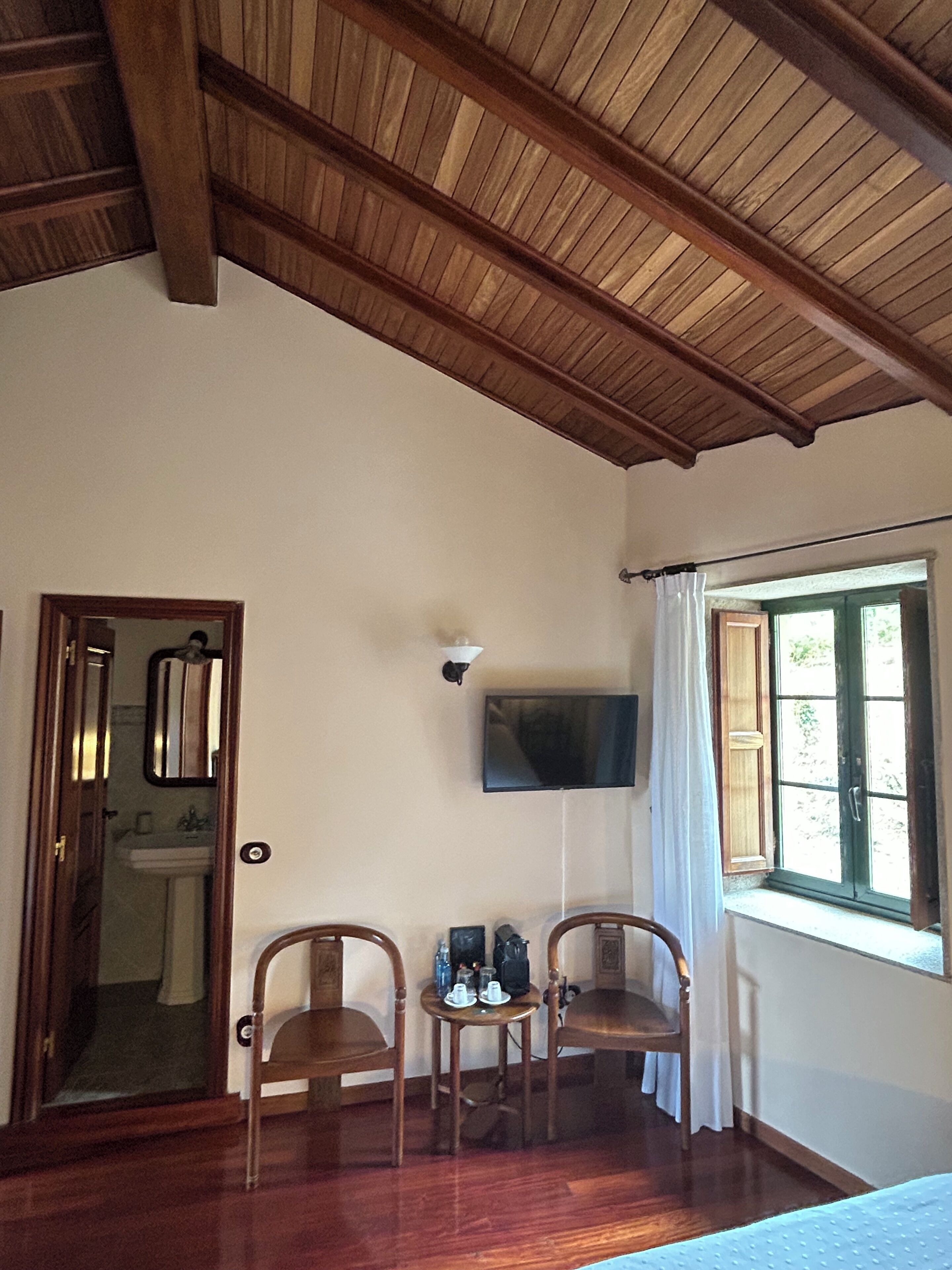 Una habitación con vigas de madera en el techo, un televisor montado en la pared, dos sillas de madera, una mesita con objetos encima y una ventana con cortinas.