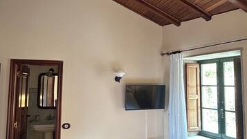 Una habitación con vigas de madera en el techo, un televisor montado en la pared, dos sillas de madera, una mesita con objetos encima y una ventana con cortinas.
