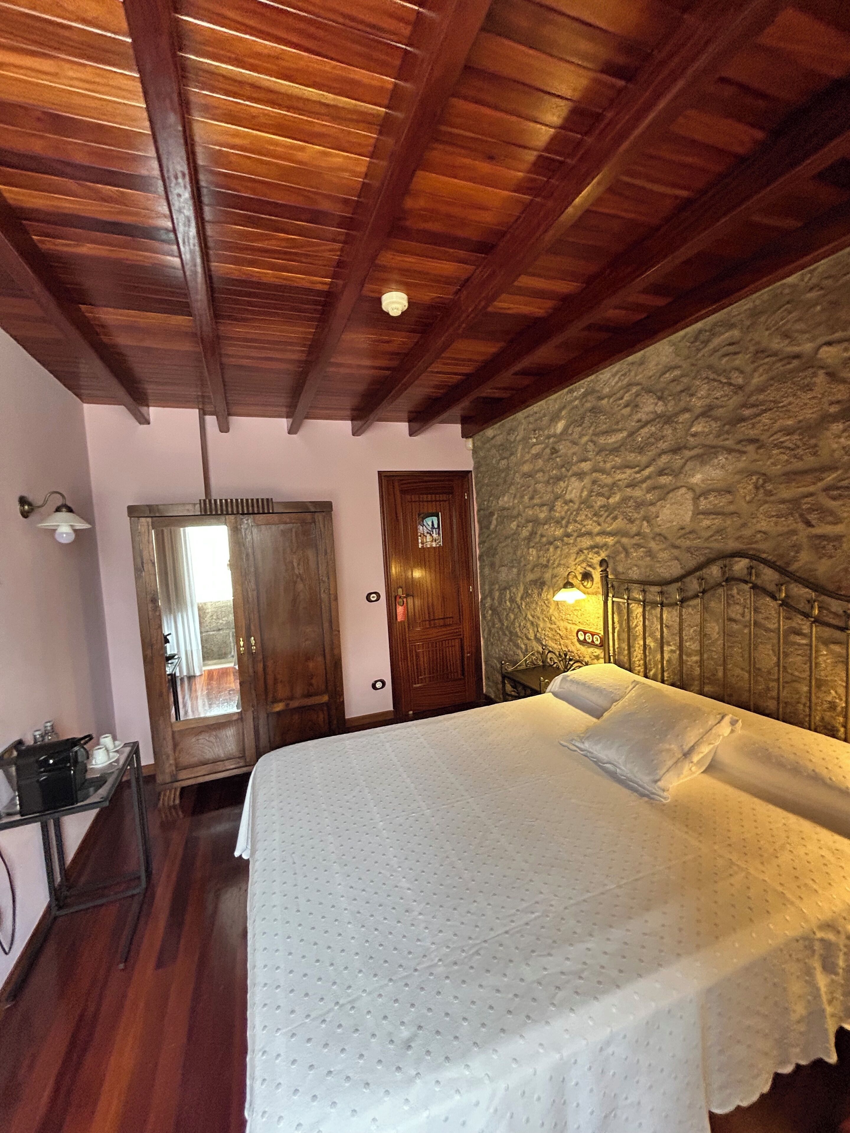 A bedroom with a wooden ceiling, a stone wall, a bed with white bedding, a nightstand, and a door leading to a balcony.