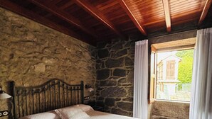 A bedroom with a stone wall, wooden ceiling beams, a bed with white bedding, and a view of a building through a window. - CASA DE COSTOIA (Ames)