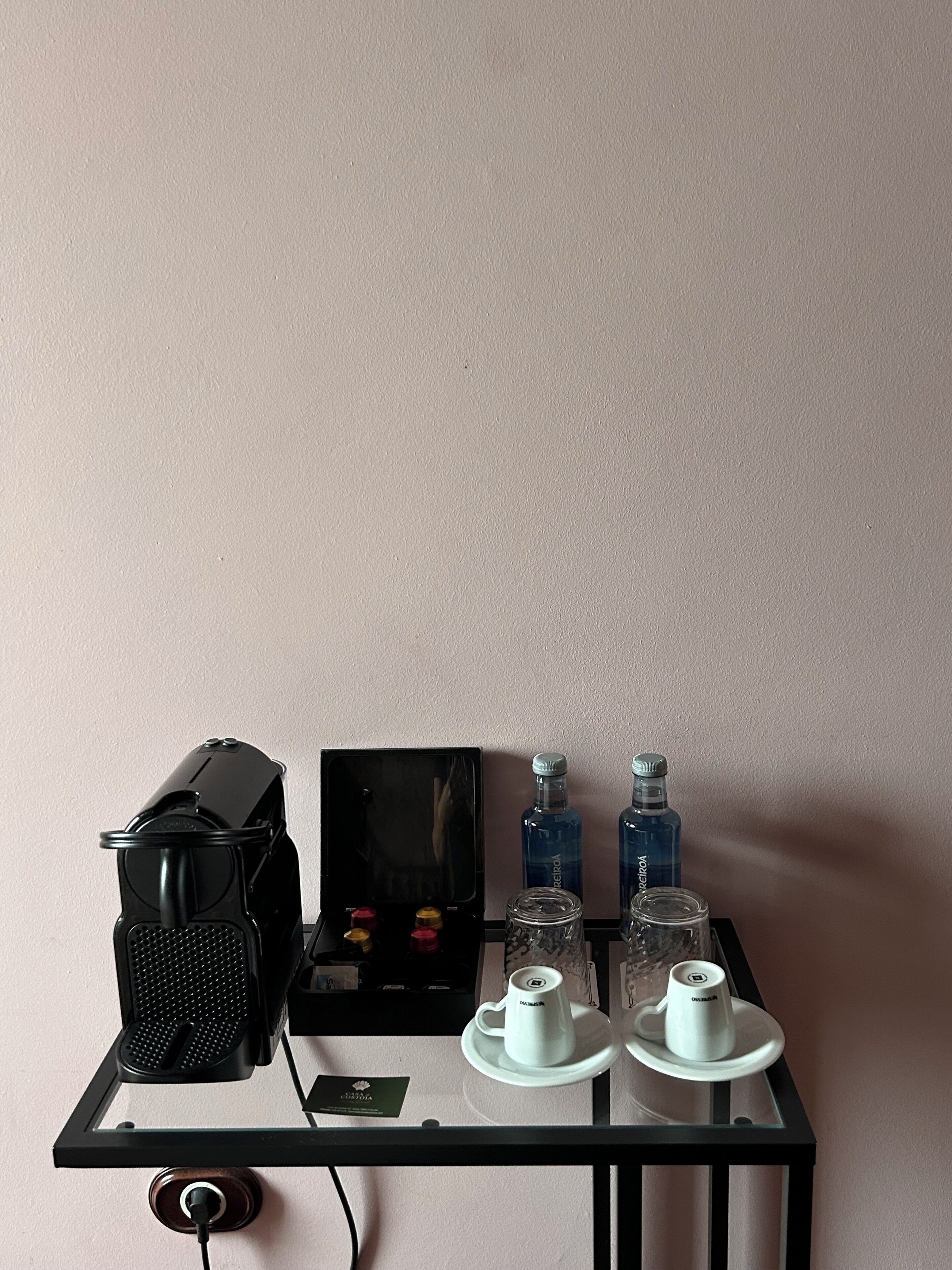 Une pièce avec une table en verre sur laquelle se trouvent un haut-parleur noir, un petit appareil noir, deux bouteilles d’eau et une théière avec une tasse.