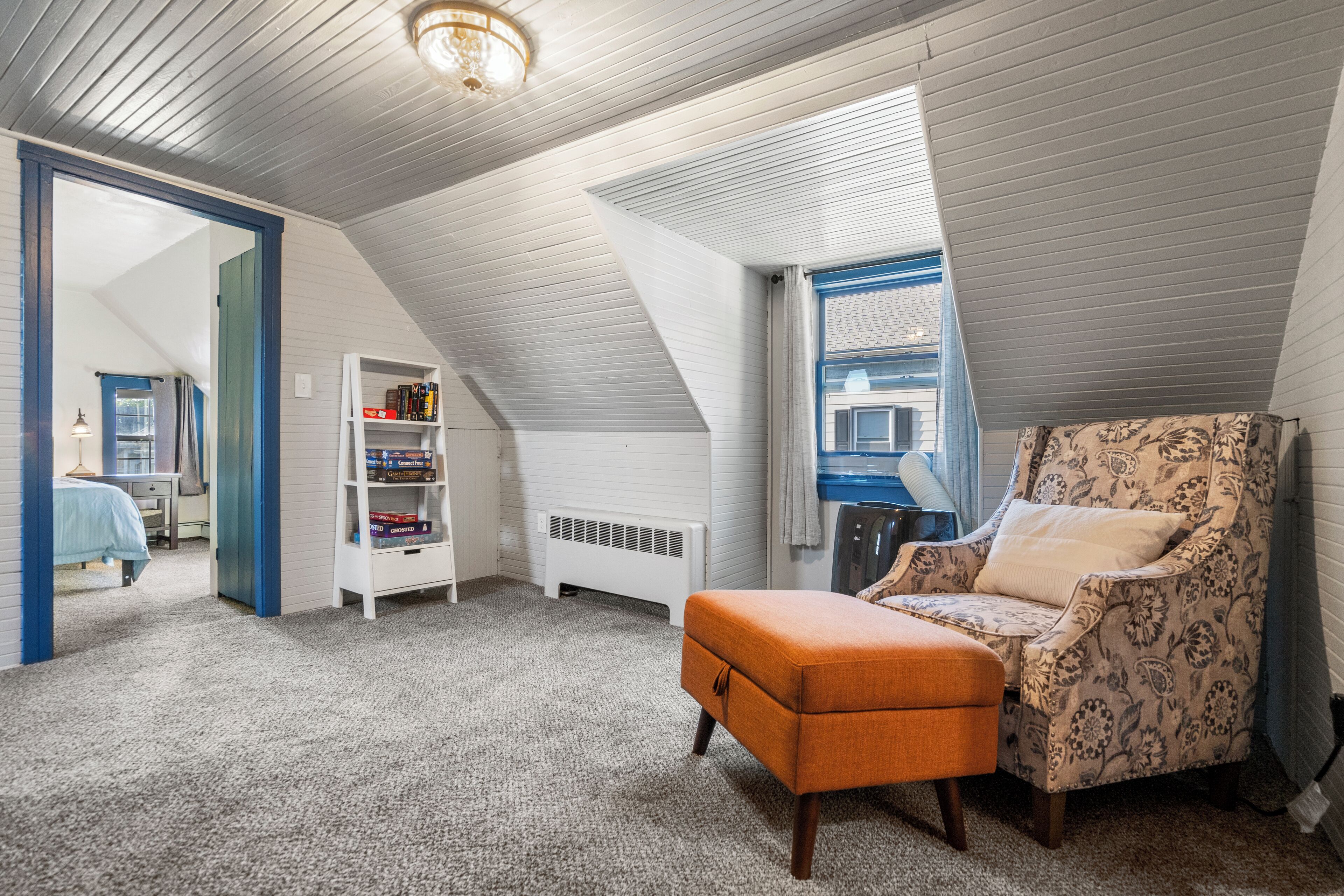 A cozy attic bedroom with a sloped ceiling, a bookshelf, a patterned armchair, and a window with curtains.