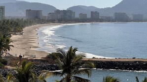 A coastal beach with a rocky shoreline, palm trees, and buildings in the background. - Spacious villa in Ixtapa with beautiful ocean views (Ixtapa Zihuatanejo)