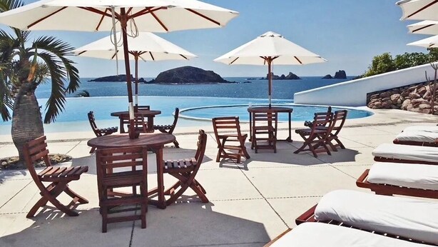 A poolside area with wooden tables and chairs, white umbrellas, and lounge chairs with white cushions. - Spacious villa in Ixtapa with beautiful ocean views (Ixtapa Zihuatanejo)