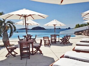 A poolside area with wooden tables and chairs, white umbrellas, and lounge chairs with white cushions.