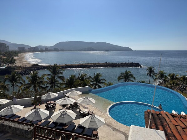 A resort pool with lounge chairs and umbrellas, a beach with palm trees, and a view of the ocean and mountains.