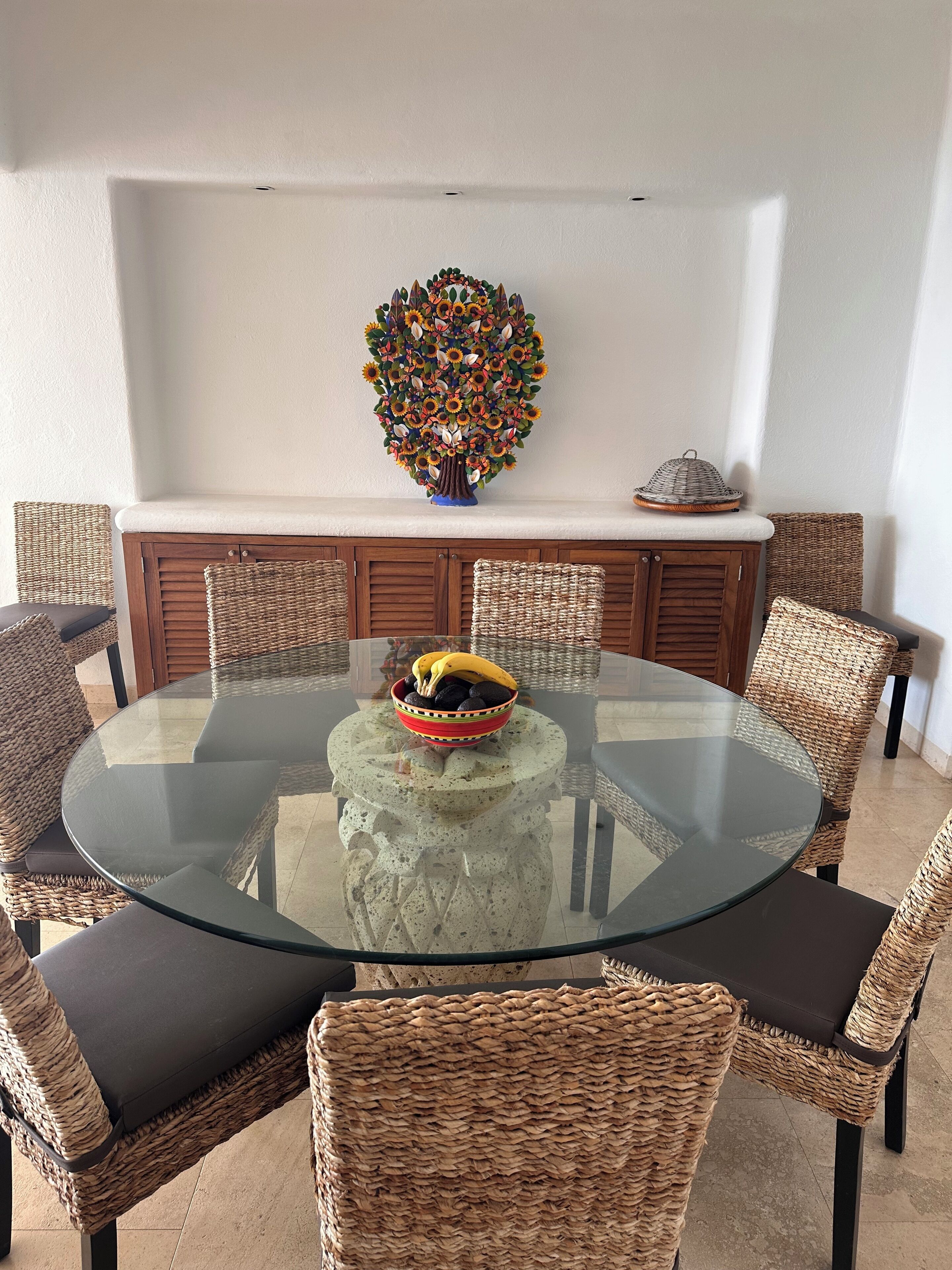 A round glass table surrounded by wicker chairs with cushions, a decorative wall piece above, and a wicker basket with fruits on the table.