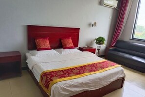 Une chambre d’hôtel avec un grand lit, une tête de lit rouge, deux tables de chevet, une petite plante, un canapé gris et une fenêtre avec des rideaux.