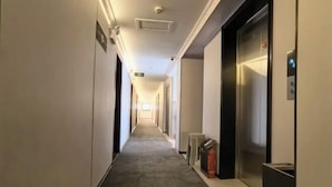 A long, narrow hotel corridor with a carpeted floor, a fire extinguisher, and an elevator on the right side. - Jingjiang Hotel (Linhai)