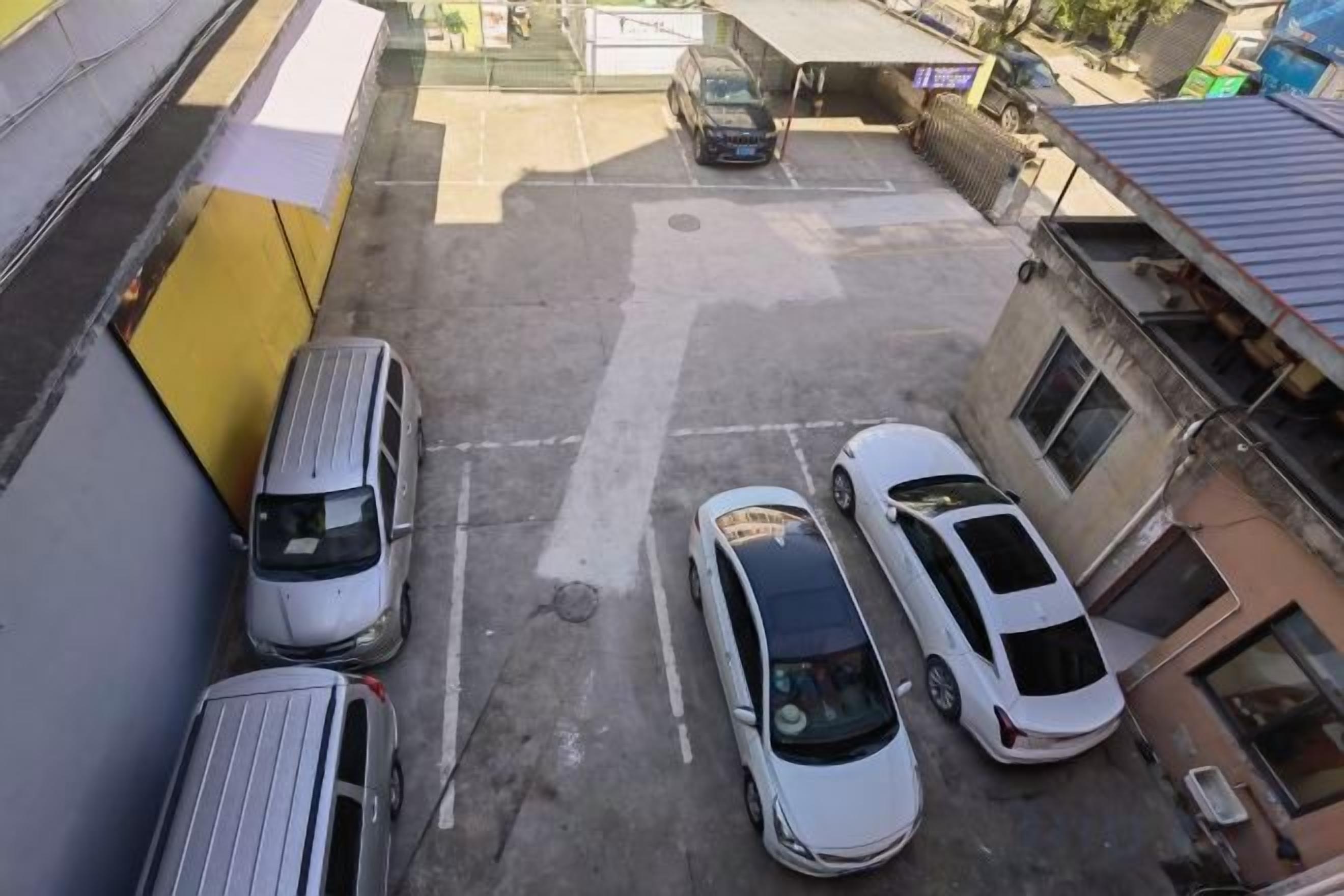 Un estacionamiento con varios coches aparcados, un edificio con pared amarilla y una estructura pequeña al fondo.