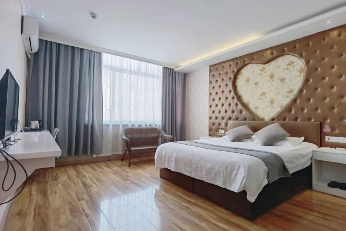 Ein Hotelzimmer mit einem großen Bett, einem herzförmigen Kopfteil, einem Schreibtisch mit Stuhl und einem Fenster mit Vorhängen.