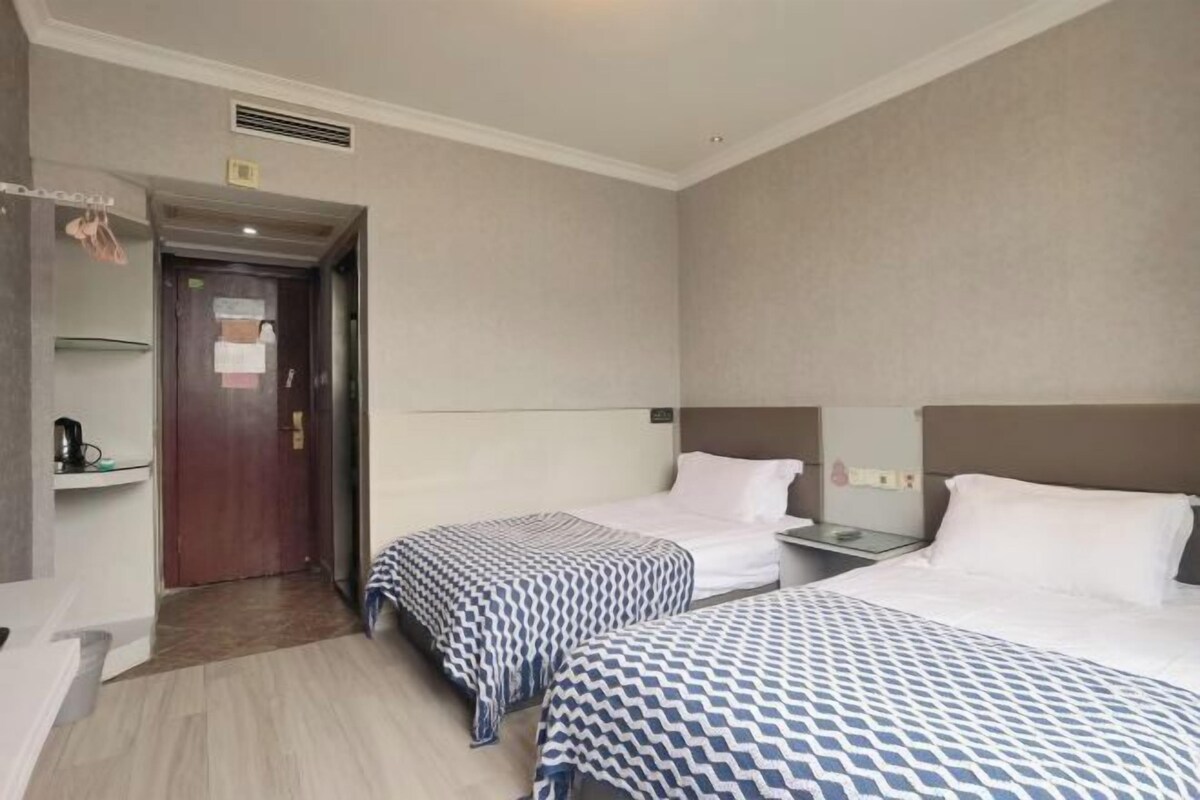 A hotel room with two beds, a wooden door, a small table, and a shelf with a kettle.
