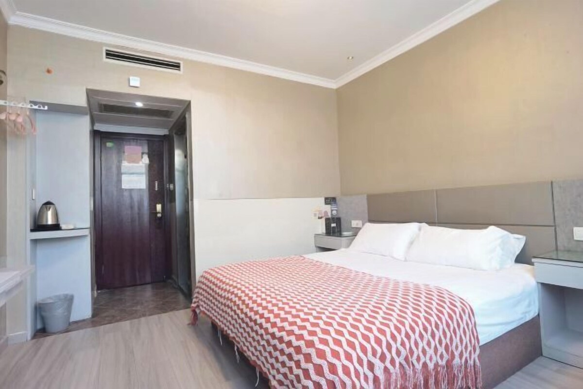 A hotel room with a bed, bedside table, and a small desk with a chair. There is a door leading to another room and a trash bin near the door.