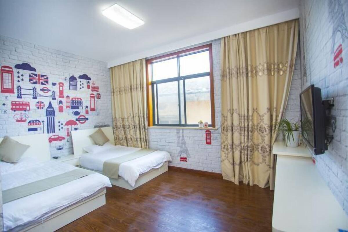 A hotel room with two beds, a large window with curtains, a brick-patterned wall, and a television on a stand.