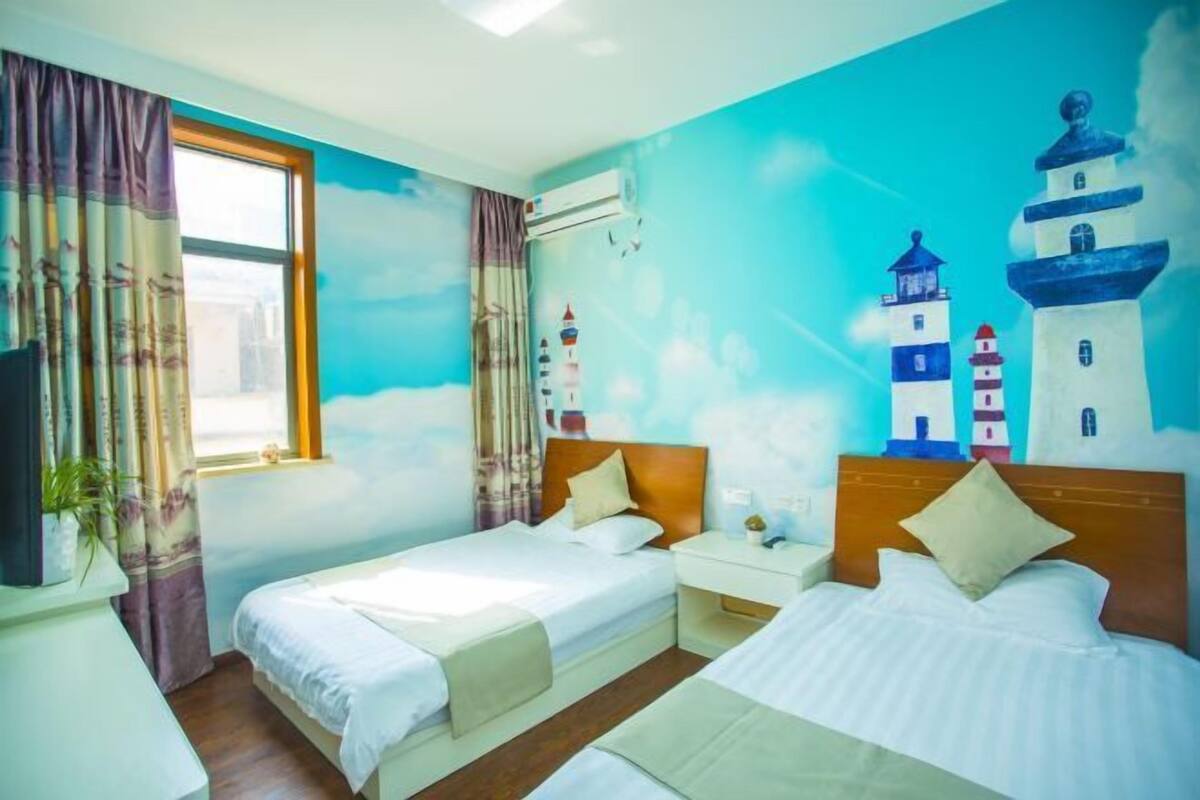 A room with two beds, a window with curtains, a wall mural of lighthouses, and a small table with a plant.