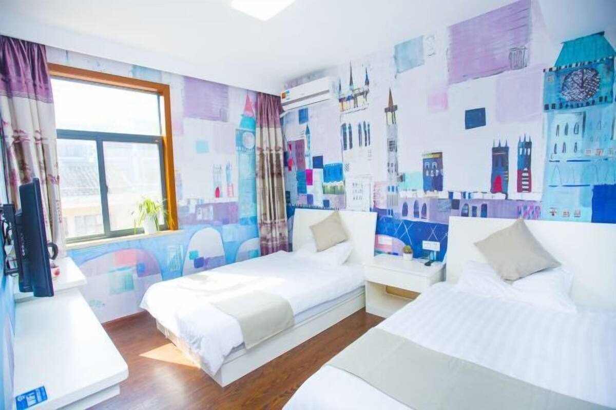 A hotel room with two beds, a large window, and a wall decorated with cityscape murals.