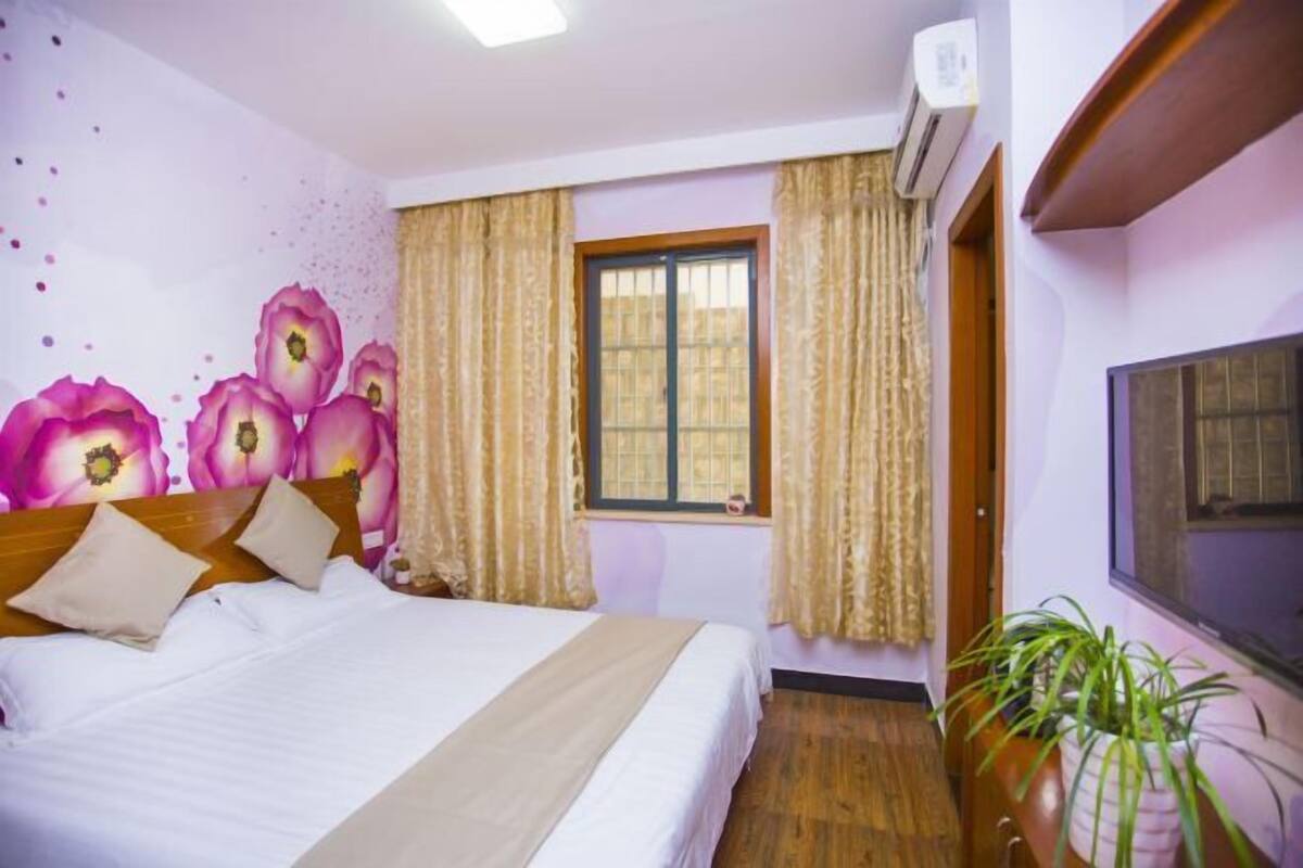 A hotel room with a bed, curtains, a window, an air conditioning unit, and a potted plant.
