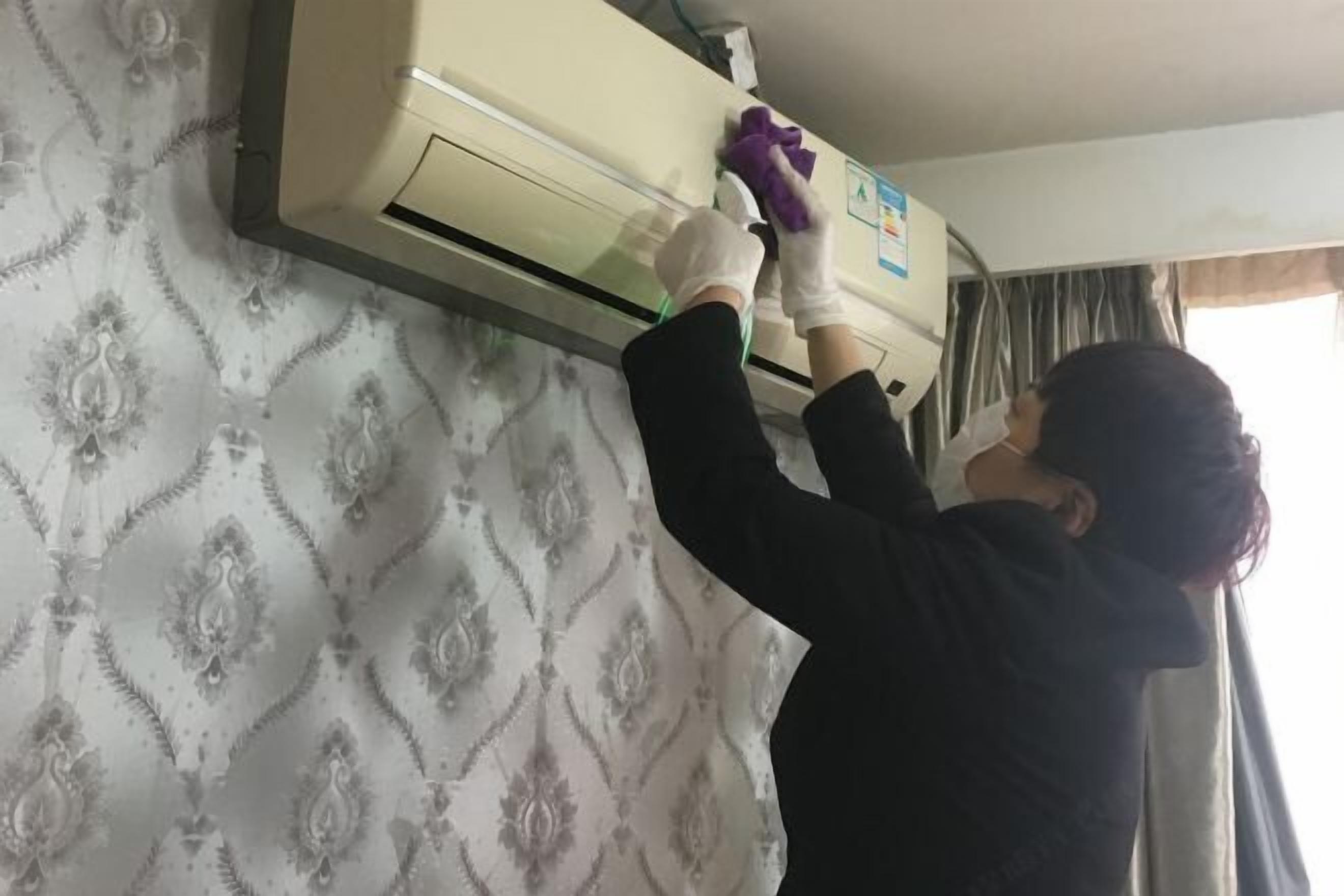 A person cleaning an air conditioning unit on a wall with patterned wallpaper.