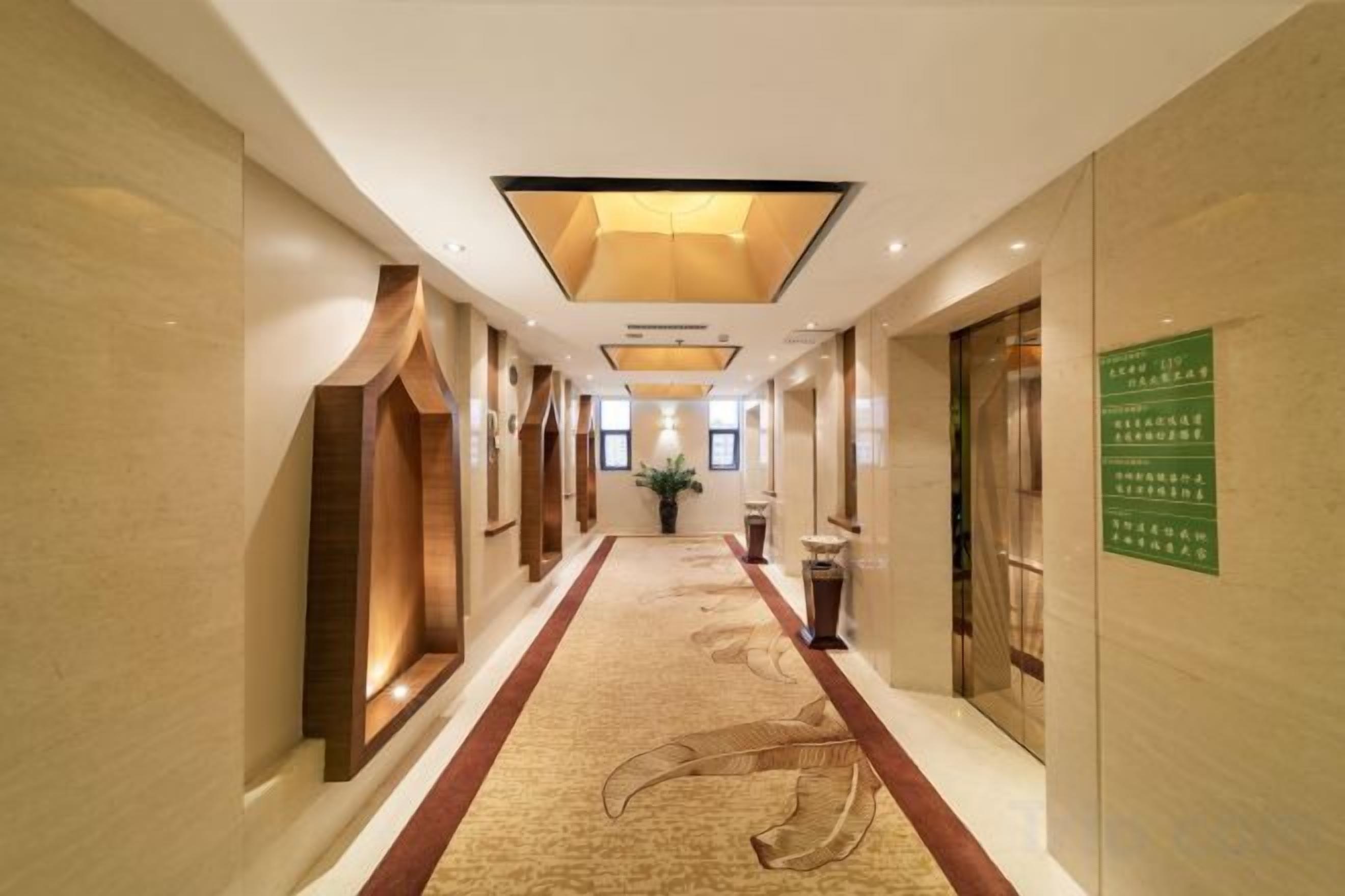 A hotel corridor with a carpeted floor, recessed lighting, and a green sign on the wall.