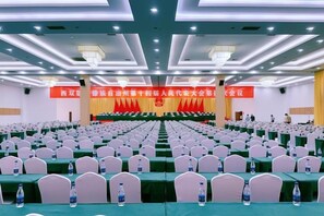 A conference room with rows of tables and chairs, a stage with a podium, and banners on the walls. - State lnvestment Exhibition Hotel (Jinghong)