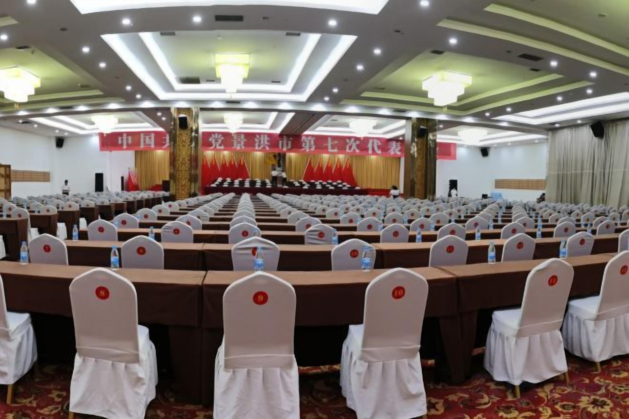 A conference room with rows of tables and chairs, a stage with a banner, and a chandelier.