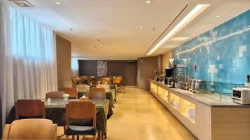 A hotel dining area with tables and chairs, a counter with a coffee machine, and a blue-tiled wall.